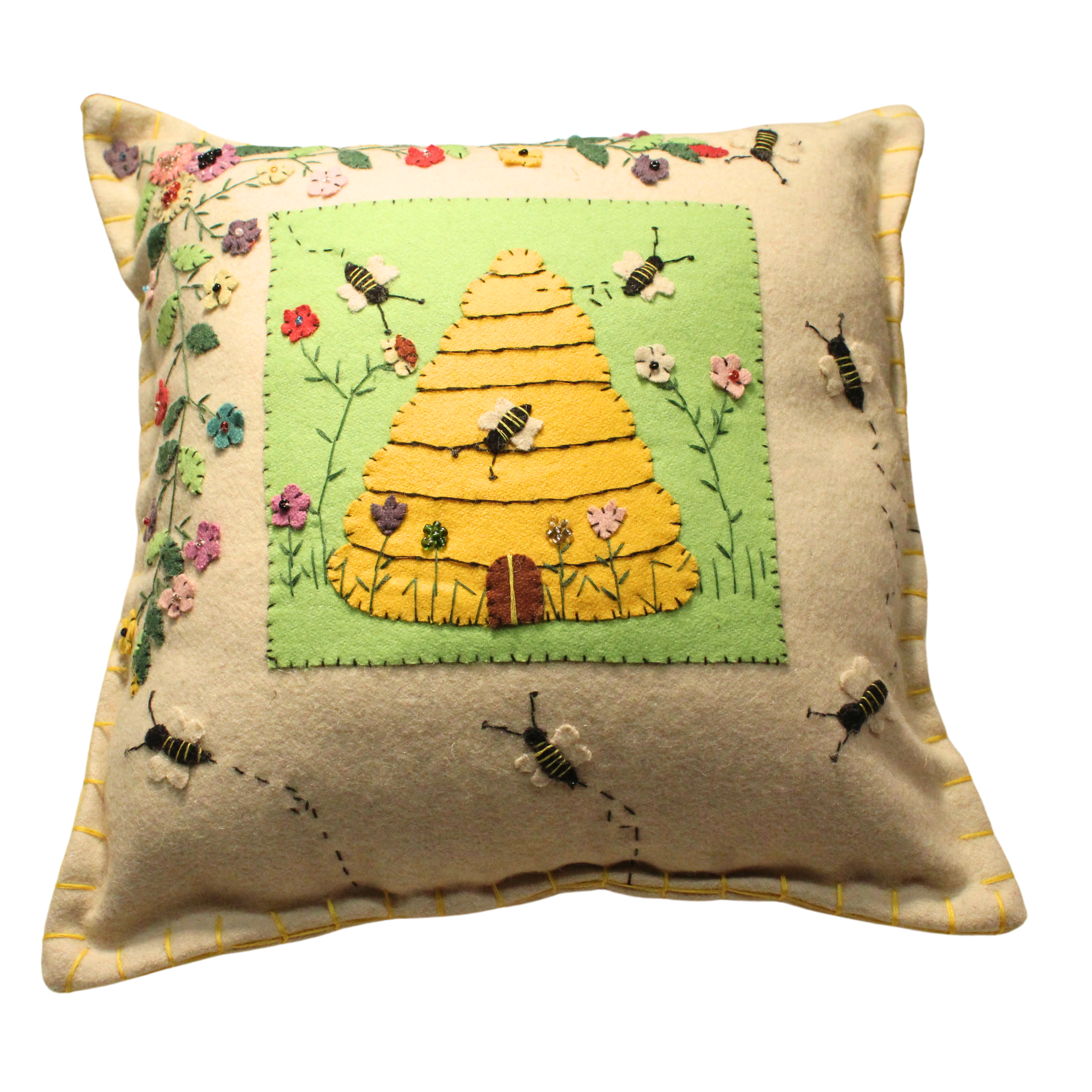 A square beige fragrance pillow with a yellow beehive on a green patch of grass. There are several bees flying around the beehive and flowers. The pillow has a yellow stitched border.