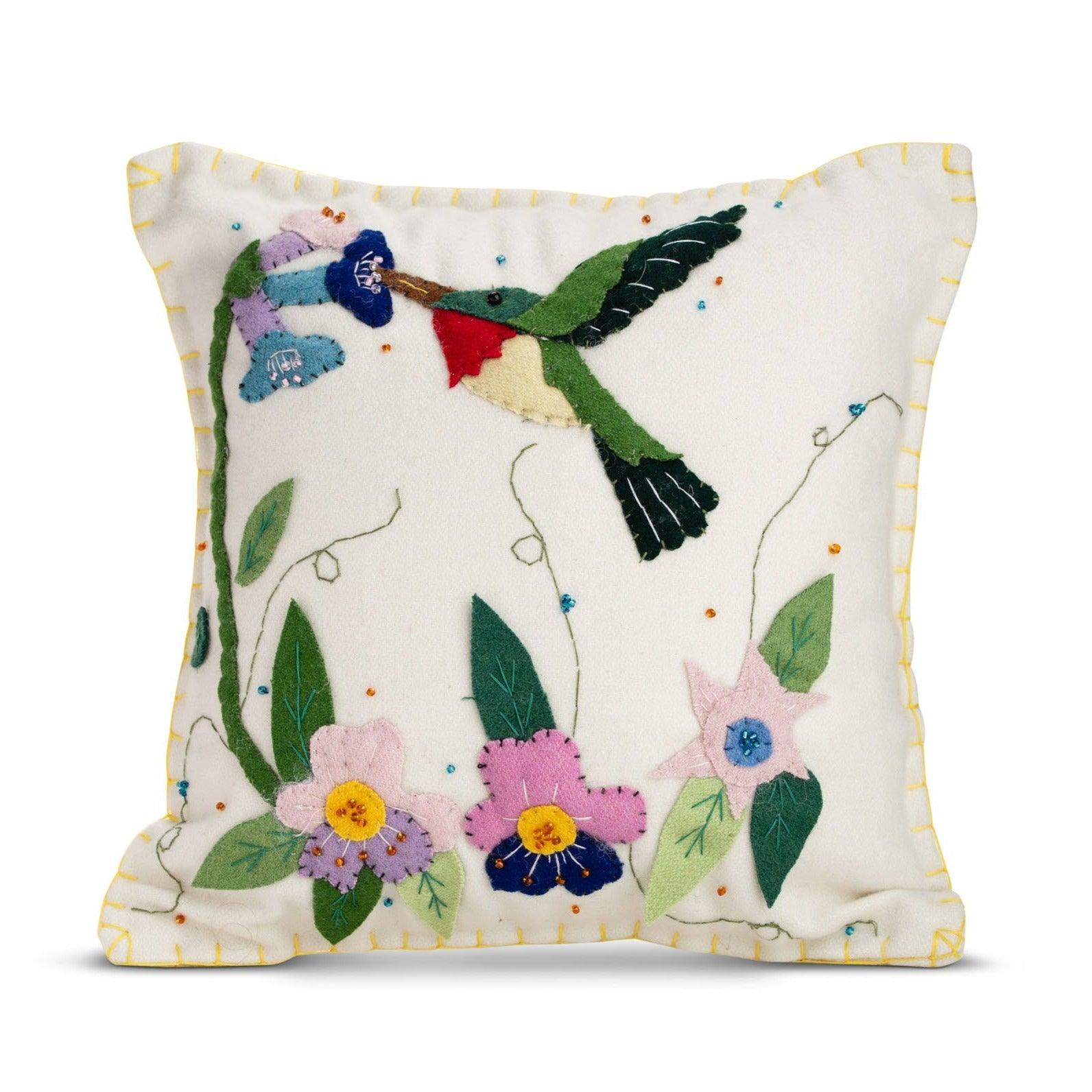 A square felt beige pillow with a colorful hummingbird hovering over pink and purple flowers and green leaves. The hummingbird has a green body, red throat, and black wings. The pillow has a yellow stitched border.