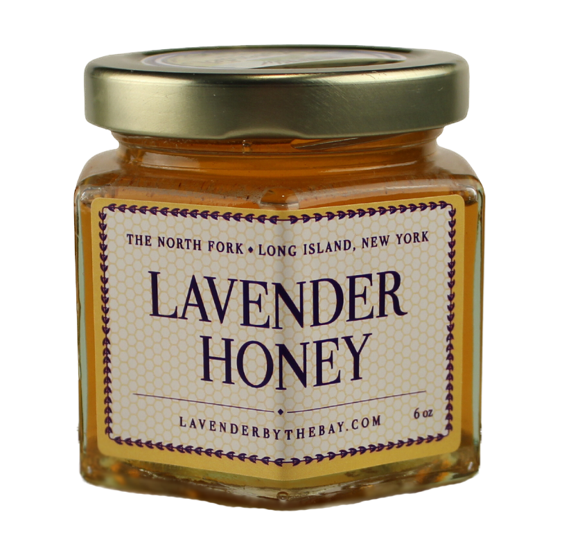 A hexagonal glass jar filled with golden raw honey, with a gold lid. The jar has a label reading "Lavender Honey" and the location "The North Fork, Long Island, New York