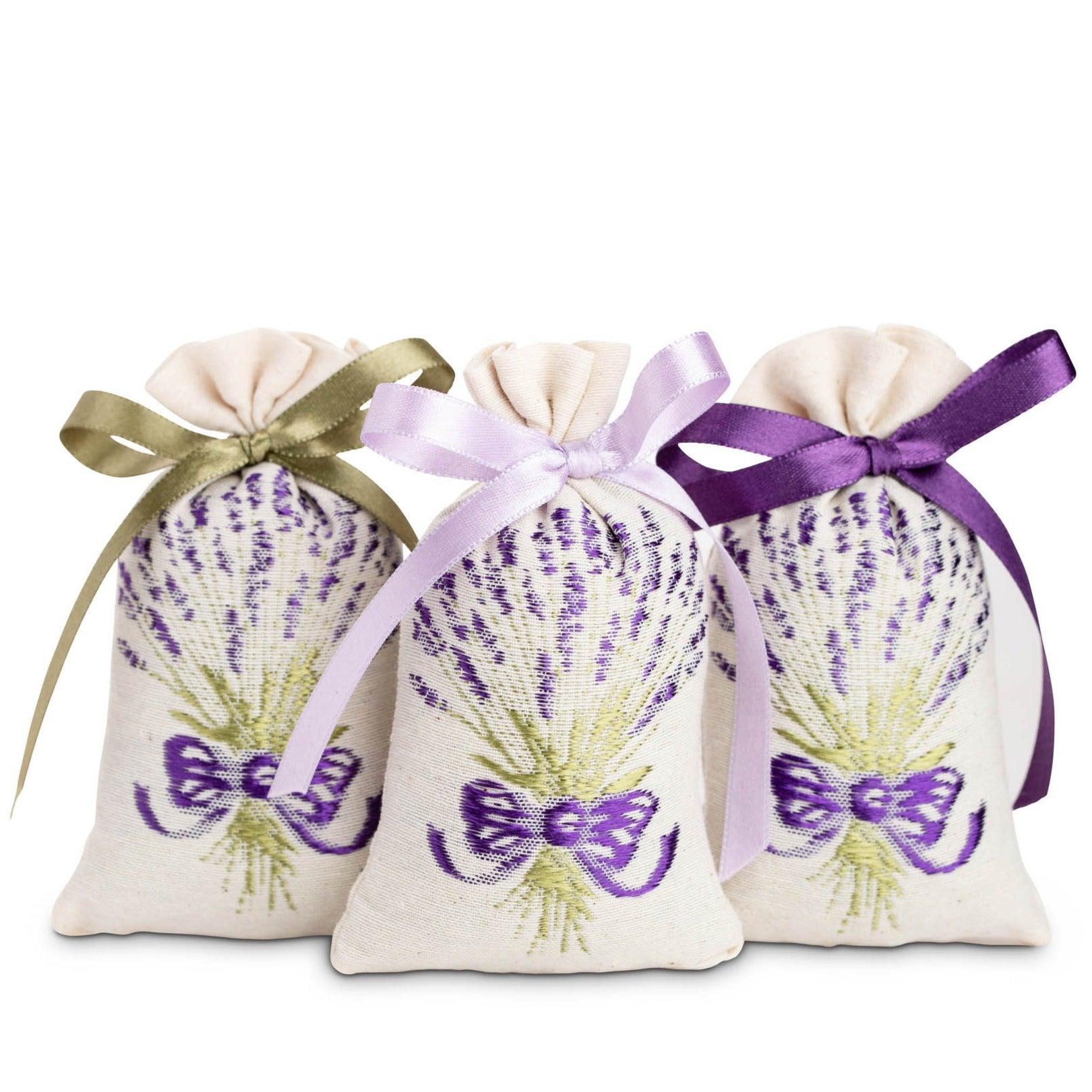 The image appears to show three small lavender sachets with a lavender plant design embroidered on the front. Each bag is tied with a ribbon in a different color: purple, green, and lavender. The sachet bags are filled with dried lavender.