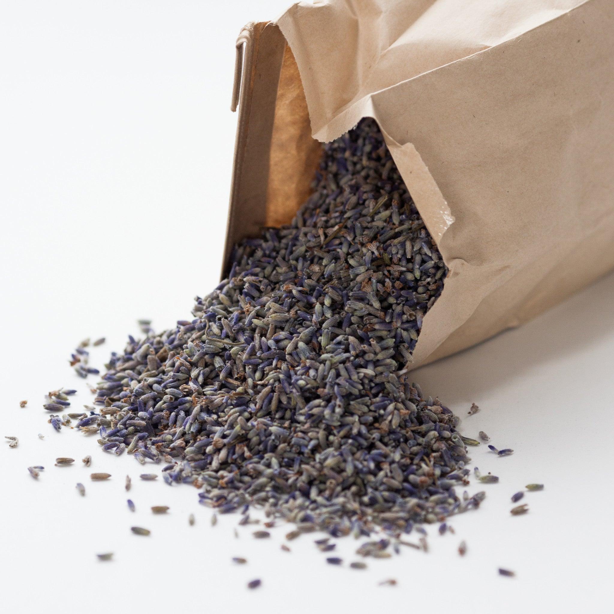 A brown paper bag with loose  lavender spilling out onto a white surface. The lavender buds are light purple color.
