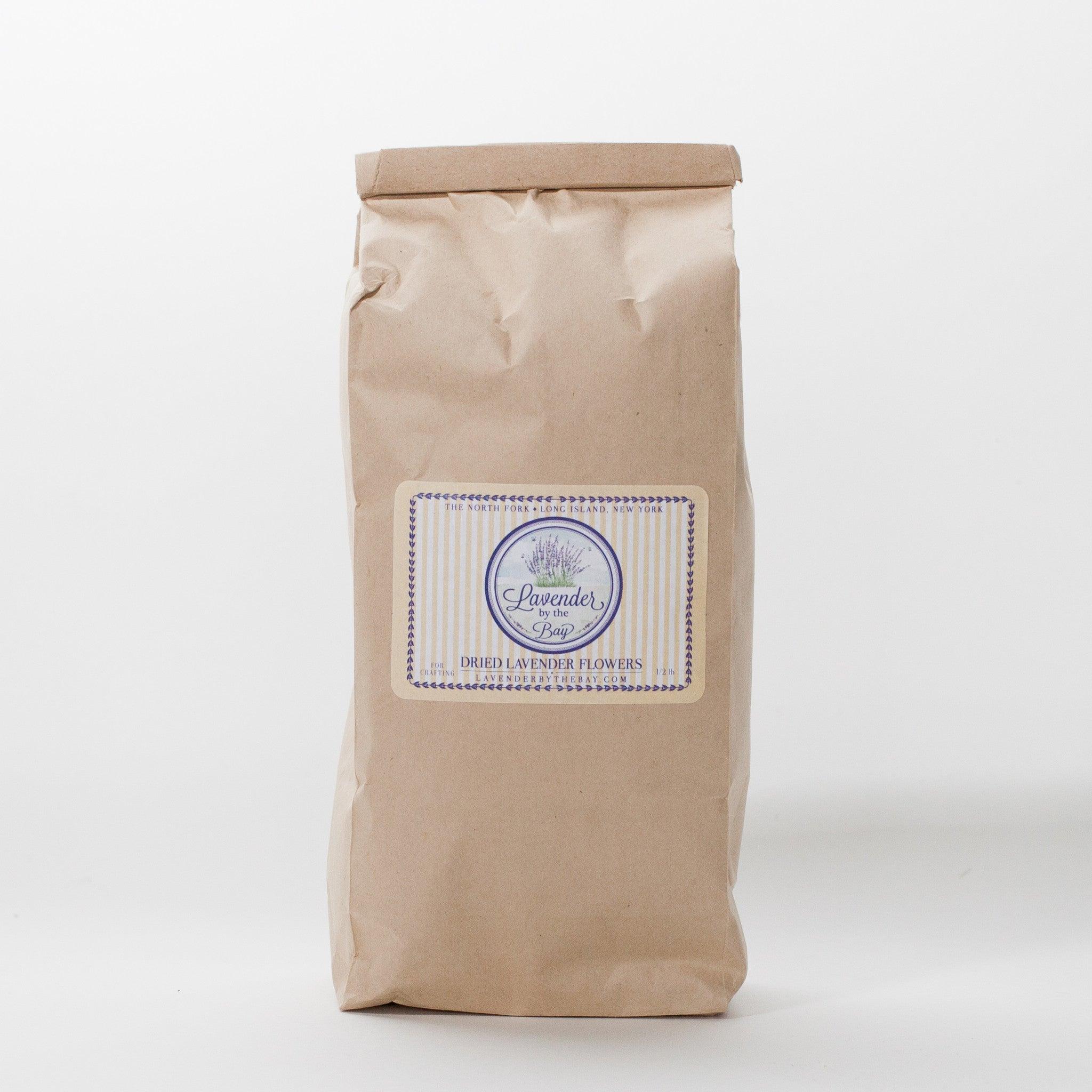 A closed brown paper bag filled with dried French lavender flowers. The bag has a label that says "Lavender by the Bay" and "Dried Lavender Flowers.