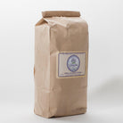 A closed brown paper bag filled with dried French loose lavender buds. The bag has a label that says "Lavender by the Bay" and "Dried Lavender Flowers. shot taken  at an angle