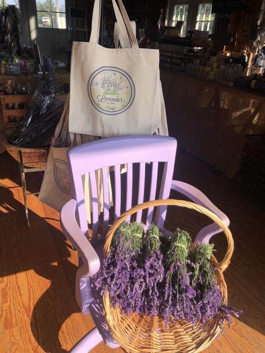 canvas tote bags with handprinted the Lavender By The Bay logo hanged on a rack behined a blue chair with lavender flowers in a wooden floor store