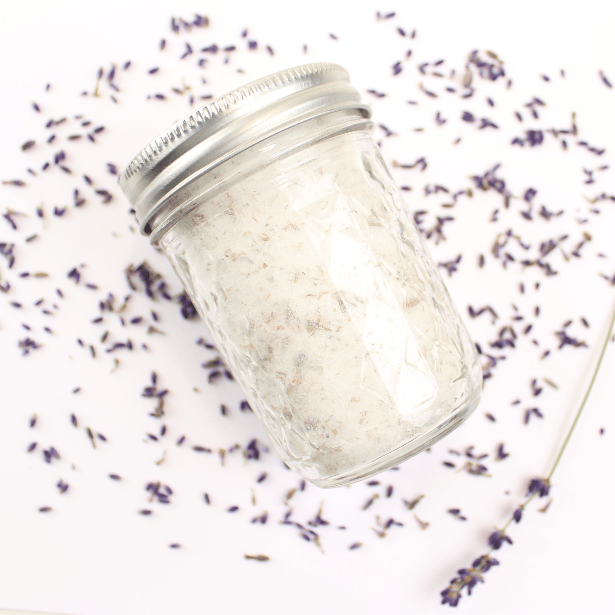 A glass jar filled with Lavender Sugar, surrounded by dried lavender flowers.