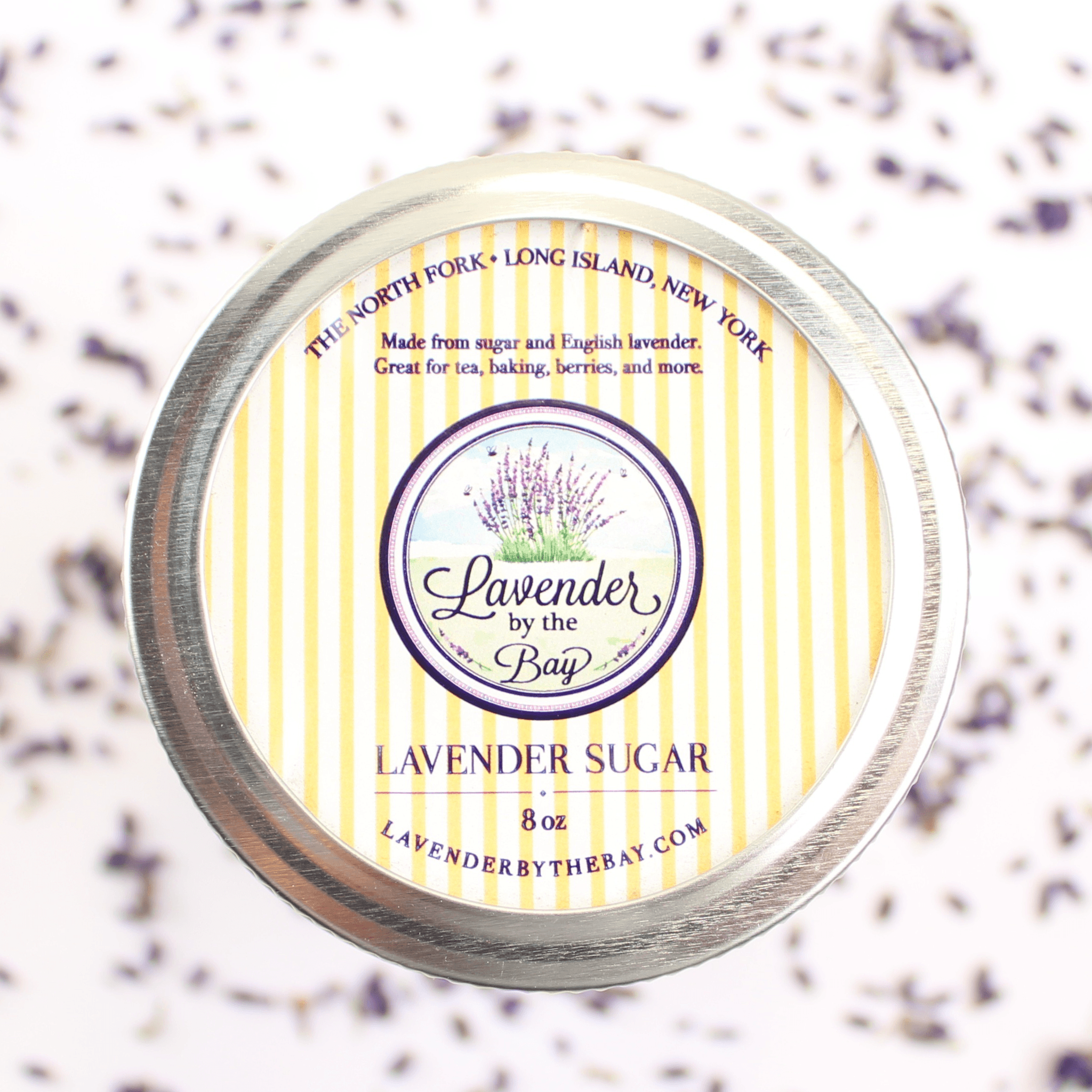 A round metal jar filled with lavender sugar, with a colorful label that includes the name "Lavender by the Bay" and information "made from sugar and english lavender great for tea, baking, barries and more"