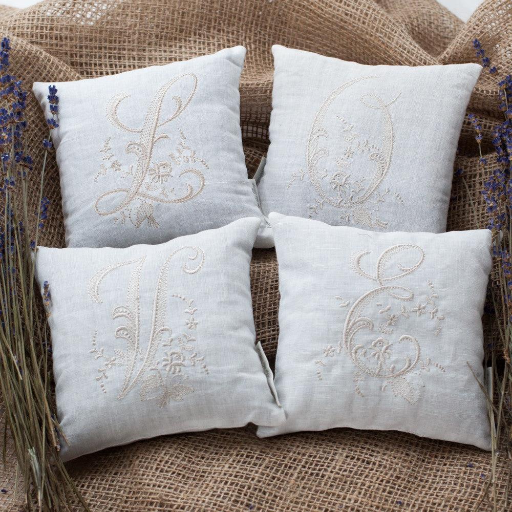 4 white Monogrammed pillows each pillow features a different embroidered letters L Q V E in a similar style The pillows are arranged on a burlap background with lavenders flowers bouquet on each side