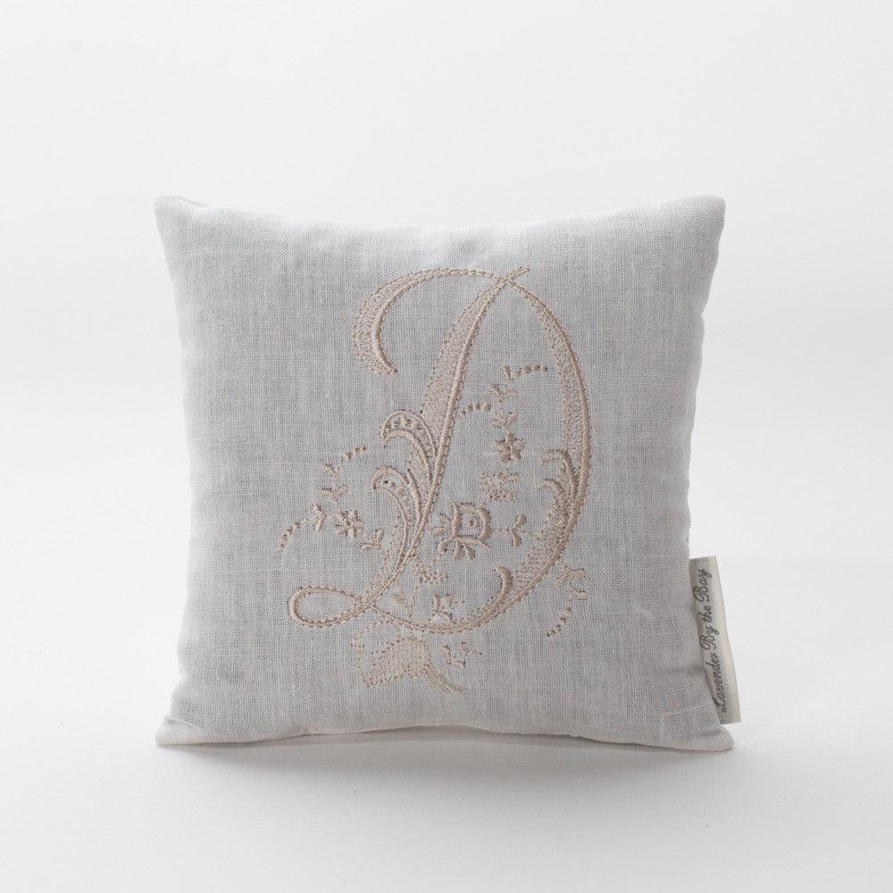 The image depicts a beautifully square, off-white embroidered monogram pillow with the letter "D," intricately stitched with delicate floral patterns and swirls. The pillow appears soft and luxurious.