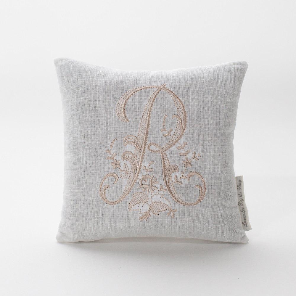 A luxurious monogram bed pillow. The pillow features a beautifully embroidered monogram of the letter "R," adorned with delicate floral patterns and swirling flourishes. The intricate stitching and elegant design make this pillow a timeless addition to any bedroom décor.