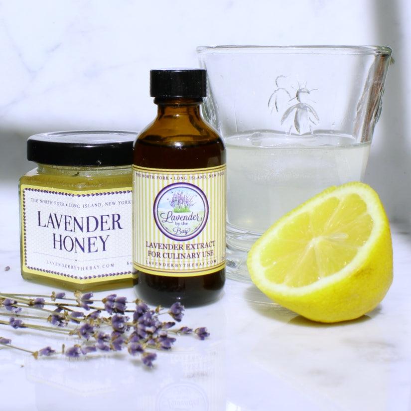 Lavender Honey Lemonade featured image showing a jar of lavender honey, a bottle of lavender extract, a glass of water, and a sliced lemon on a white surface