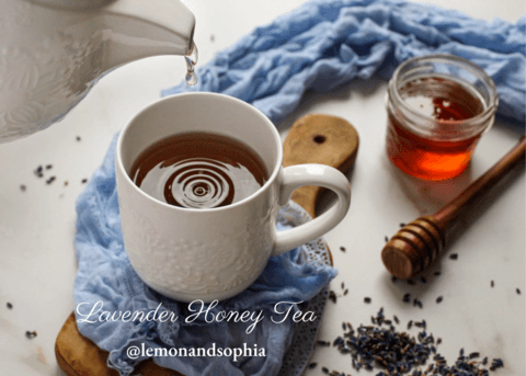 Lavender Tea Time Recipe featured image showing A white mug of lavender honey tea being poured from a teapot. A jar of honey, a honey dipper, and dried lavender flowers are scattered around the mug on a wooden board Lemon and Sophia