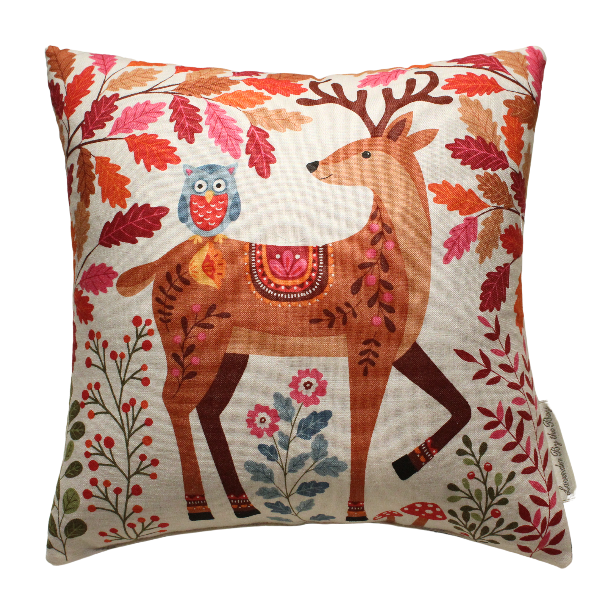 A square pillow with a cream-colored background features a brown deer with a colorful geometric pattern on its back. An orange owl sits on the deer's back, looking towards the left. The background is filled with pink and red flowers, leaves, and berries.